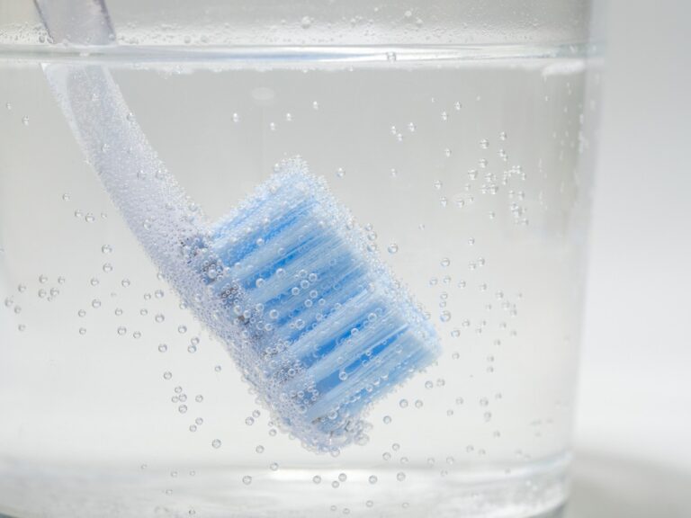 A manual toothbrush bristles down in a glass of bubbly disinfectant.