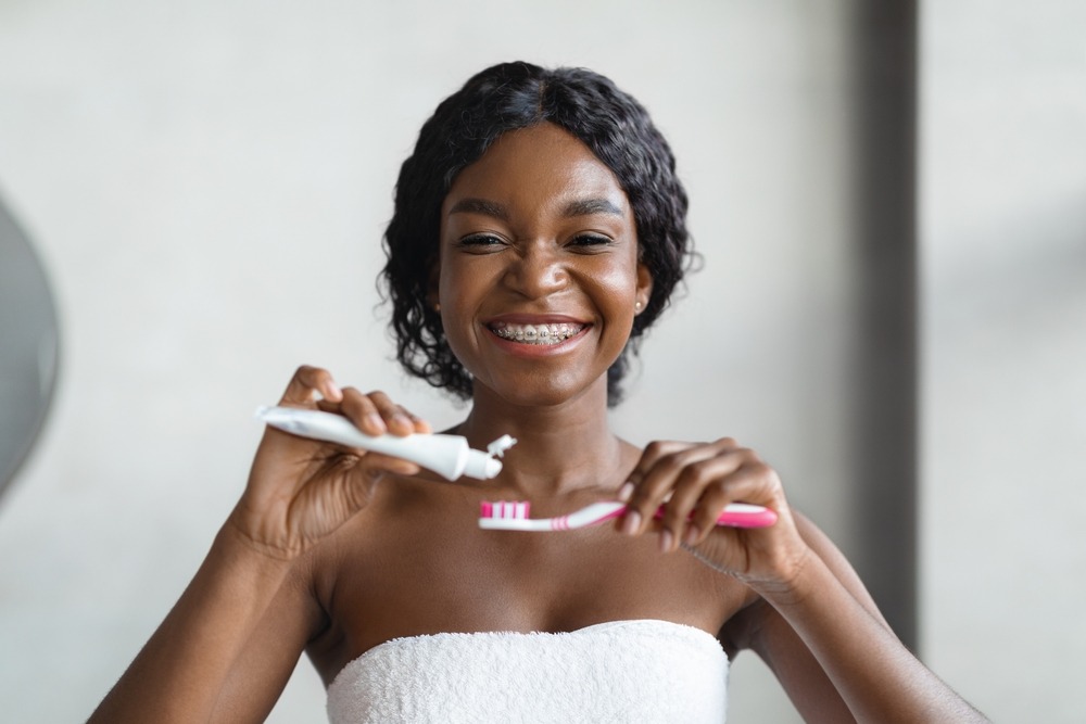 A smiling woman with braces applies toothpaste to a manual toothbrush.