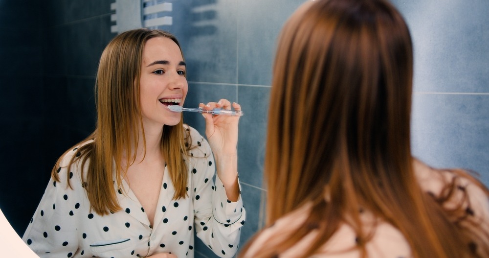 A teen with braces looks in the mirror while brushing her teeth with a manual toothbrush.